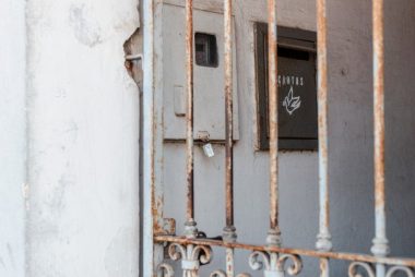 A rusty metal fence frames an old mail drop against a worn wall, creating a rustic atmosphere.